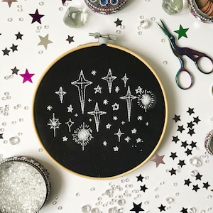 Pu&ograve; includere: Un cerchio da ricamo nero con ricamo a filo bianco di stelle su sfondo nero. Il cerchio &egrave; circondato da coriandoli a forma di stella argentati, neri e colorati e perline di vetro trasparenti.