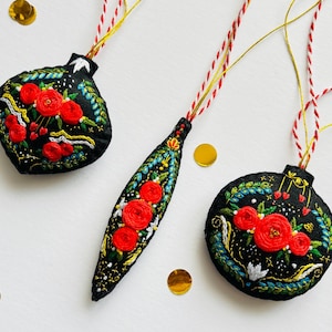 Może przedstawiać: Trzy czarne ozdoby filcowe z czerwonymi haftowanymi kwiatami i zielonymi liśćmi. Ozdoby wiszą na czerwonej i złotej sznurku.