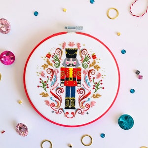 Puede incluir: Arte de bordado en aro con un cascanueces colorido con abrigo rojo y pantalones azules, rodeado de diseños florales y espirales.