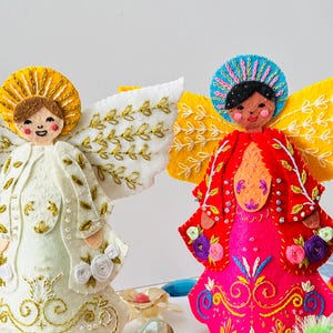 Puede incluir: Dos adornos de ángel de fieltro, uno con alas blancas y un halo dorado, el otro con alas amarillas y un halo azul. Ambos ángeles llevan vestidos bordados de colores.