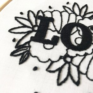 May include: Black embroidery of the word "LOVE" surrounded by floral designs on a white fabric background.