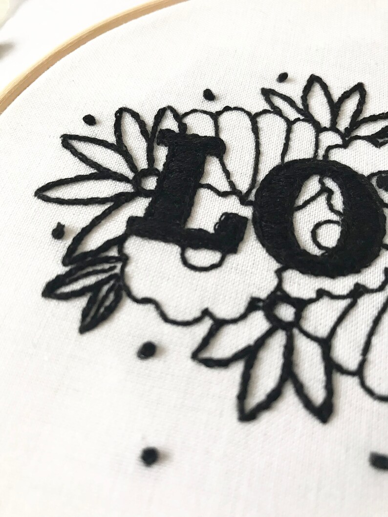 May include: Black embroidery of the word "LOVE" surrounded by floral designs on a white fabric background.