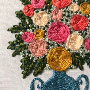 May include: A close-up of a hand-stitched embroidery of a bouquet of flowers in a blue vase. The flowers are pink, white, and yellow and are arranged in a circular pattern. The vase has a blue and white design.