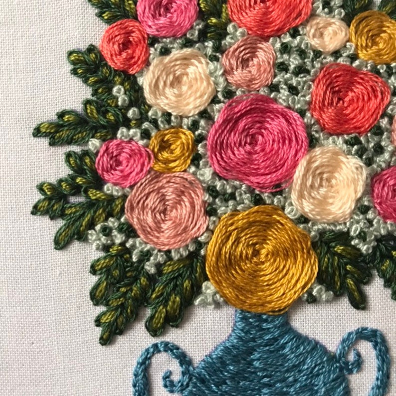 May include: A close-up of a hand-stitched embroidery of a bouquet of flowers in a blue vase. The flowers are pink, white, and yellow and are arranged in a circular pattern. The vase has a blue and white design.
