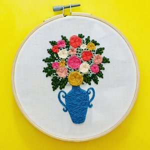 May include: Embroidered hoop art featuring a blue vase with a bouquet of pink, yellow, and white flowers.