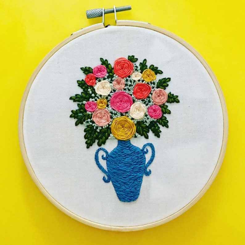 May include: Embroidered hoop art featuring a blue vase with a bouquet of pink, yellow, and white flowers.