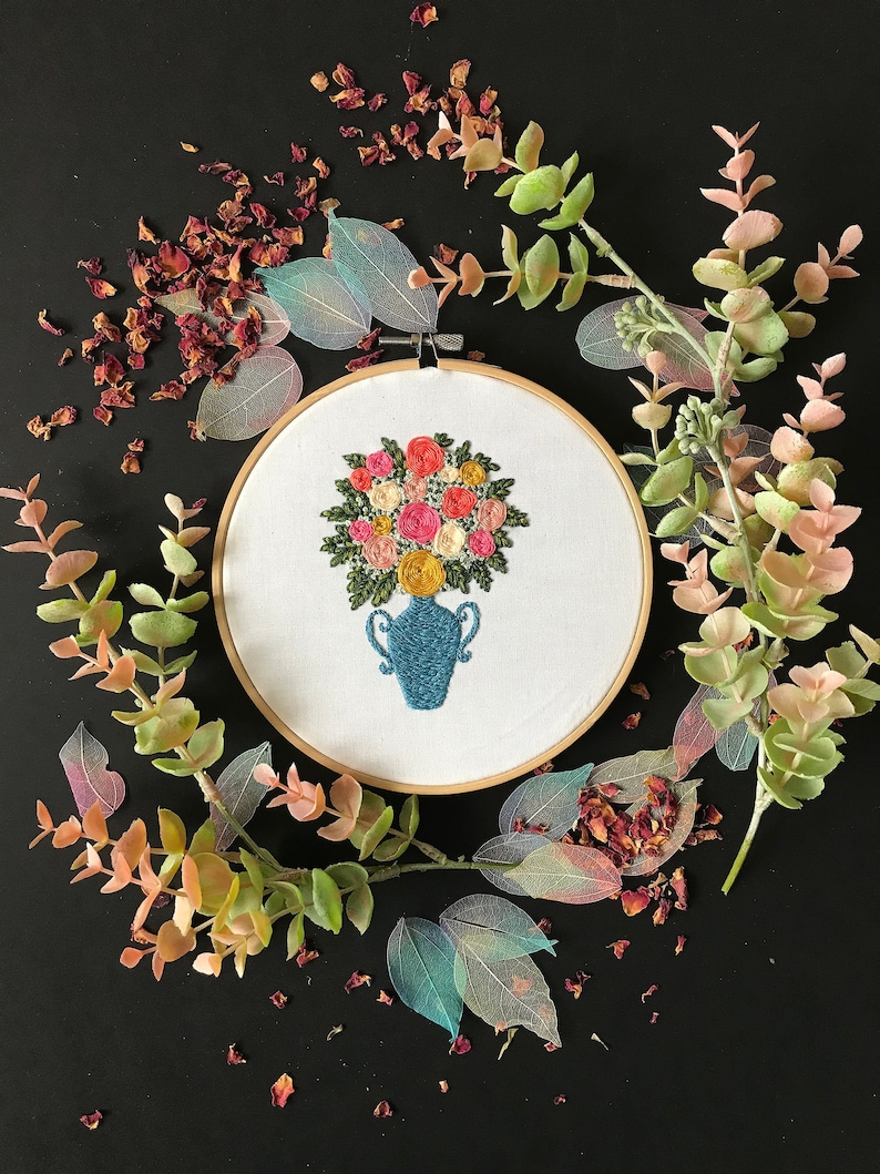 May include: Embroidered floral bouquet in a blue vase, surrounded by eucalyptus branches and dried rose petals on a black background.