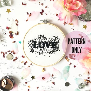 May include: A white embroidery hoop with black embroidery of the word "LOVE" surrounded by floral designs. The hoop is on a white background with pink and white flower petals, gold stars, and small white beads.