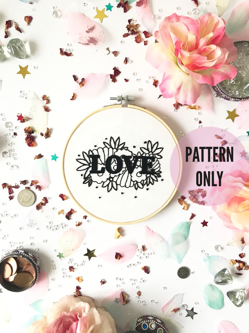 May include: A white embroidery hoop with black embroidery of the word "LOVE" surrounded by floral designs. The hoop is on a white background with pink and white flower petals, gold stars, and small white beads.