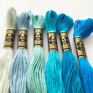 Puede incluir: Seis madejas de hilo de bordar DMC en tonos azules y verdes. Cada madeja está etiquetada con "M 25" y "8 m (8.7 yd)". Los colores varían del azul claro al turquesa intenso, ideales para bordado y punto de cruz.
