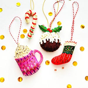 Puede incluir: Cuatro adornos navideños de fieltro que incluyen una taza rosa con nata, un bastón de caramelo, un pudin marrón con glaseado blanco y acebo, y un calcetín rojo. Cada adorno presenta bordados detallados y un cordel para colgar.