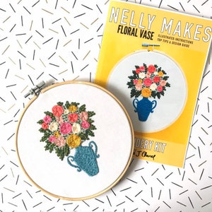 May include: A close-up of a completed embroidery hoop with a floral design. The hoop is white with a wooden frame. The embroidery depicts a blue vase filled with pink, yellow, and white flowers. The embroidery kit is titled "Nelly Makes Floral Vase" and includes illustrated instructions and design tips.