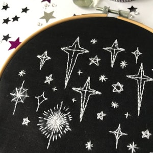 Pu&ograve; includere: Un cerchio da ricamo nero con ricamo a filo bianco di stelle e un fuoco d'artificio su uno sfondo di tessuto nero.