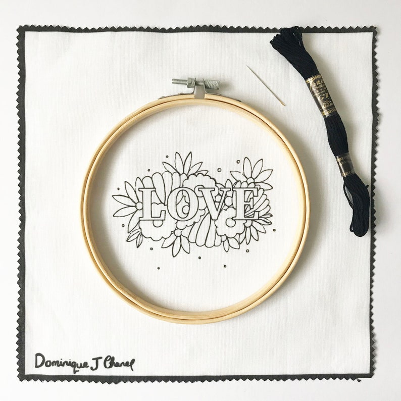 May include: A white embroidery hoop with a black outline of the word "LOVE" surrounded by flowers. The hoop is on a white fabric with a black border. A black embroidery needle and black embroidery floss are on the right side of the image.