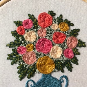 May include: A close-up of a hand-stitched embroidery design of a bouquet of pink, red, and yellow flowers in a blue vase. The flowers are arranged in a circular pattern and are surrounded by green leaves.