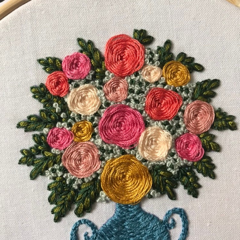 May include: A close-up of a hand-stitched embroidery design of a bouquet of pink, red, and yellow flowers in a blue vase. The flowers are arranged in a circular pattern and are surrounded by green leaves.