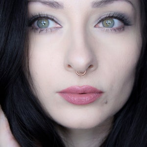 Può includere: Primo piano di un viso con un piercing al setto nasale. La persona ha i capelli scuri, gli occhi verdi e indossa del trucco.