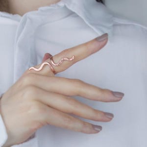 Może przedstawiać: Miedziany pierścionek o falistym, wężowym wzorze. Pierścionek jest noszony na palcu, a na zdjęciu widoczna jest dłoń i część białej koszuli. Unikalny kształt i kolor pierścionka są w centrum uwagi.