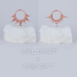 May include: A rose gold septum ring with a delicate wire design that resembles a sunburst. The ring is displayed on a white rock base. The text "VALIANT + ONE GIFT" is displayed below the ring.