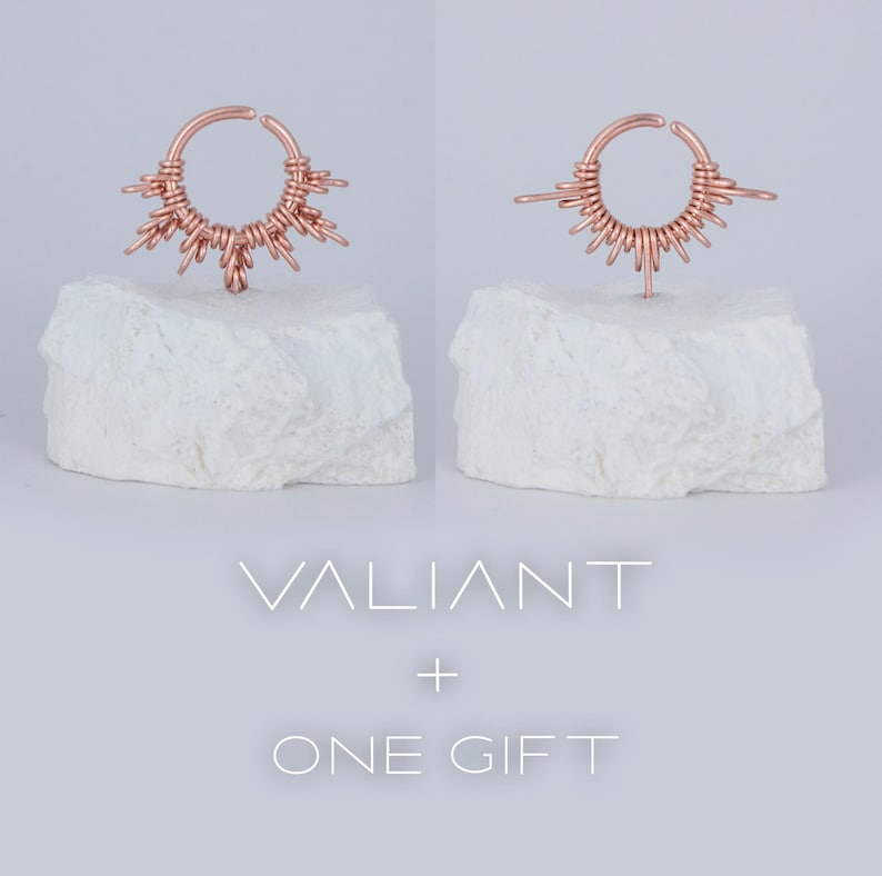 May include: A rose gold septum ring with a delicate wire design that resembles a sunburst. The ring is displayed on a white rock base. The text "VALIANT + ONE GIFT" is displayed below the ring.