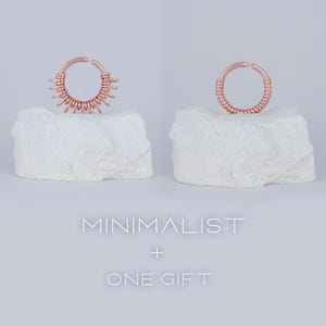 May include: Two rose gold nose rings on white rock-like surfaces. The ring on the left has a more intricate design with multiple loops. The ring on the right is a simple, continuous loop. The text "MINIMALIST + ONE GIFT" is below the rings.