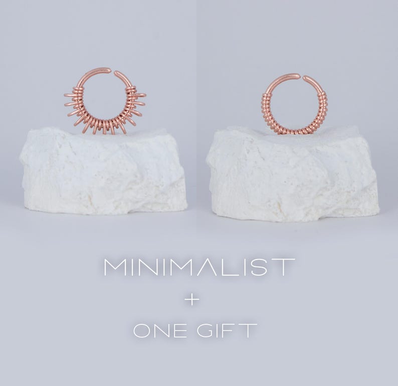 May include: Two rose gold nose rings on white rock-like surfaces. The ring on the left has a more intricate design with multiple loops. The ring on the right is a simple, continuous loop. The text "MINIMALIST + ONE GIFT" is below the rings.