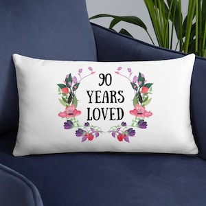 May include: White throw pillow with a floral wreath design and the text "90 Years Loved".