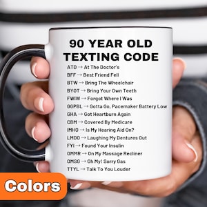 May include: A white ceramic mug with a black handle and rim, featuring the text "90 YEAR OLD TEXTING CODE" and a list of acronyms with their meanings. The mug also has the text "8 Colors" in an orange box.