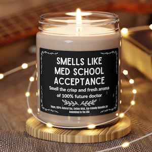 Puede incluir: Una vela marrón en un tarro de vidrio con una etiqueta negra que dice "Smells Like Med School Acceptance. Smell the crisp and fresh aroma of 100% future doctor." La etiqueta también incluye el texto "Vegan, 100% Natural Soy, Cotton Wick, Eco-Friendly Reusable Jar. Assembled in the USA."