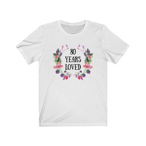 May include: White t-shirt featuring a floral wreath design. The wreath surrounds the text "80 YEARS LOVED" in black. The wreath includes pink, purple, and green watercolour-style flowers and leaves. A casual tee for a birthday celebration.