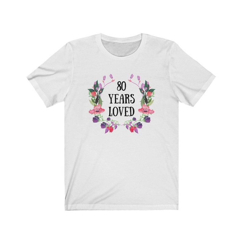 May include: White t-shirt featuring a floral wreath design. The wreath surrounds the text "80 YEARS LOVED" in black. The wreath includes pink, purple, and green watercolour-style flowers and leaves. A casual tee for a birthday celebration.