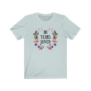 May include: A pale blue t-shirt featuring the text "80 YEARS LOVED" in black, encircled by a floral wreath with pink and purple flowers and green leaves. The shirt has a crew neck and short sleeves.