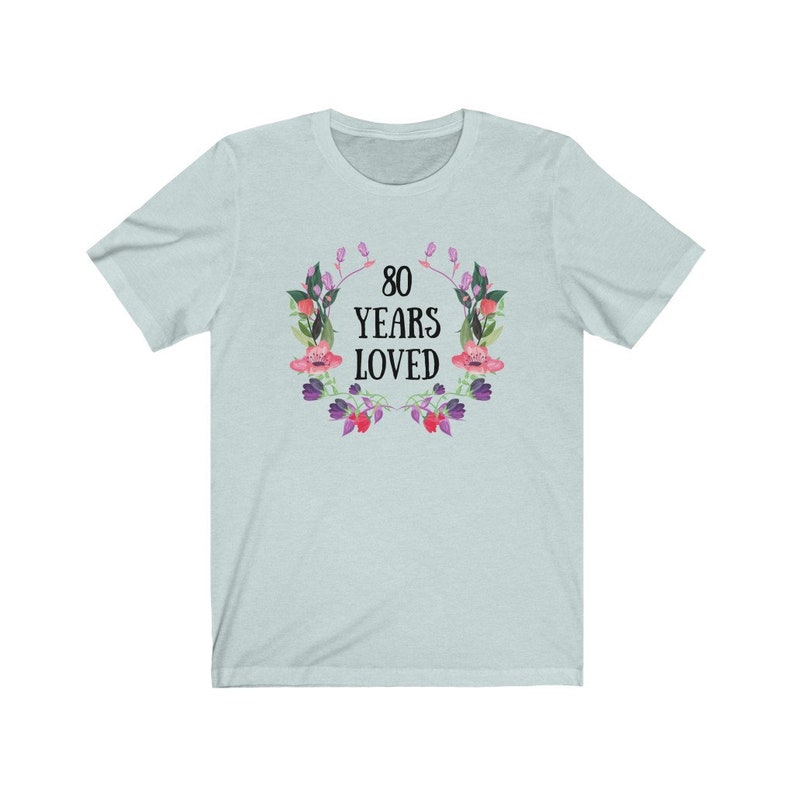 May include: A pale blue t-shirt featuring the text "80 YEARS LOVED" in black, encircled by a floral wreath with pink and purple flowers and green leaves. The shirt has a crew neck and short sleeves.