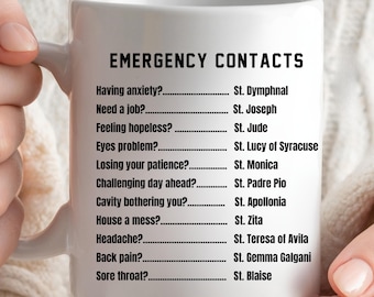 May include: White ceramic mug with black text that reads "EMERGENCY CONTACTS" and lists various situations with corresponding saints to call upon for help.
