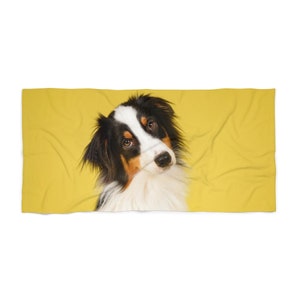 May include: A rectangular beach towel featuring a portrait of an Australian Shepherd dog with black, white, and tan fur against a solid, vibrant yellow background. The dog has a white blaze on its face and dark, expressive eyes looking forward.
