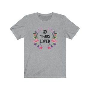 May include: A grey t-shirt with the text "80 YEARS LOVED" encircled by a floral wreath in pink, purple, and green. The short-sleeved shirt is made of a soft material.