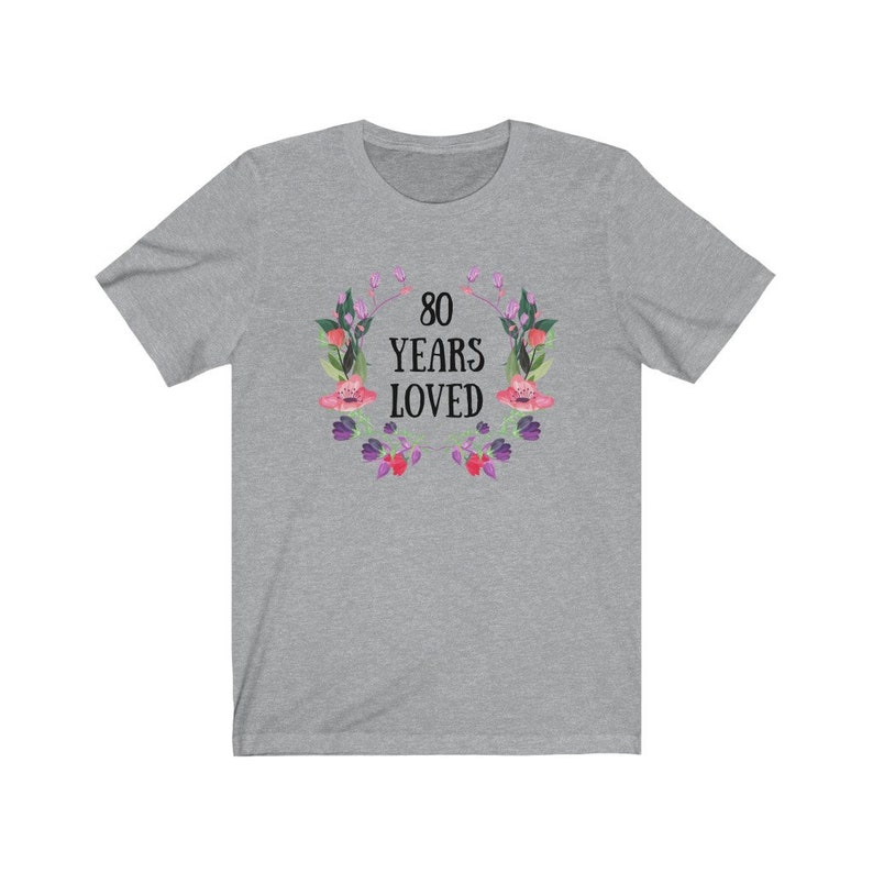 May include: A grey t-shirt with the text "80 YEARS LOVED" encircled by a floral wreath in pink, purple, and green. The short-sleeved shirt is made of a soft material.