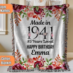 Puede incluir: Una manta de algodón tejido gris con un diseño floral en tonos rojos, rosas y verdes. La manta tiene el texto "Made in 1941", "85 Years Loved" y "Happy Birthday Emma".