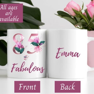 Puede incluir: Taza de cerámica blanca con un diseño floral rosa y el número 85. El texto "Fabulous" está debajo del número. La parte posterior de la taza tiene el nombre "Emma" en letras rosas.