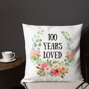 May include: White throw pillow with a floral wreath design and the text "100 Years Loved" in black.