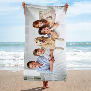 May include: A rectangular beach towel featuring a printed photograph of a family consisting of a parent, two children, and a golden retriever dog lying together on a white surface. The towel is held up against a backdrop of a sandy beach and ocean.