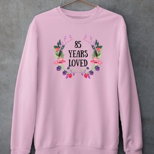 Puede incluir: Sudadera rosa con un diseño de corona floral y el texto "85 Years Loved".