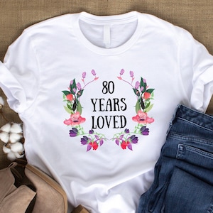 May include: White t-shirt featuring the text "80 YEARS LOVED" within a floral wreath design. The shirt is styled with blue jeans and tan shoes, on a burlap backdrop. Cotton stems are also present.