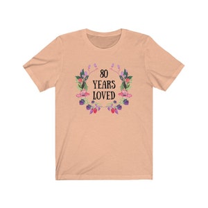 May include: A peach-coloured t-shirt with the text "80 YEARS LOVED" in black, encircled by a floral wreath with pink, purple, and green details. The shirt is made of a soft material.