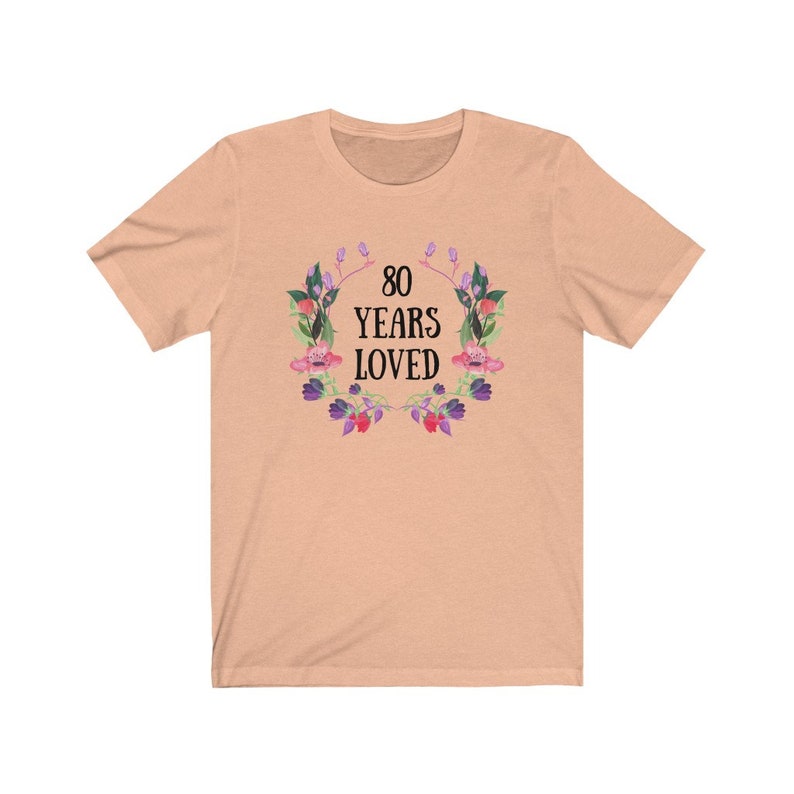 May include: A peach-coloured t-shirt with the text "80 YEARS LOVED" in black, encircled by a floral wreath with pink, purple, and green details. The shirt is made of a soft material.