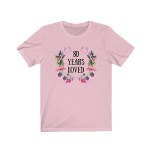 May include: A pale pink t-shirt with the text "80 YEARS LOVED" within a floral wreath. The wreath is composed of watercolour-style pink, purple, and green flowers and leaves. The shirt has a crew neck and short sleeves.