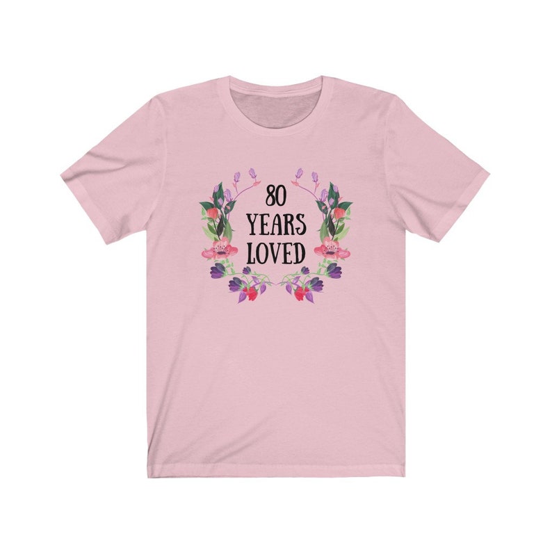 May include: A pale pink t-shirt with the text "80 YEARS LOVED" within a floral wreath. The wreath is composed of watercolour-style pink, purple, and green flowers and leaves. The shirt has a crew neck and short sleeves.
