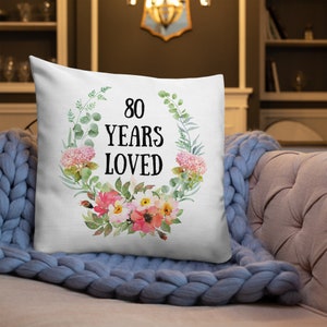 May include: White throw pillow with a floral wreath design. The wreath is made of green leaves and pink and white flowers. The text "80 Years Loved" is in the center of the wreath.