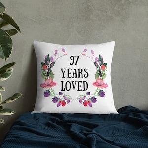 Può includere: Un cuscino bianco con un design a corona di fiori. La corona è composta da fiori rosa e viola e foglie verdi. Il testo "97 Years Loved" è al centro della corona.