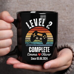 May include: Black coffee mug with a retro-style design featuring a video game controller and the text "Level 2 Complete Emma Oliver Since 05.06.2024".